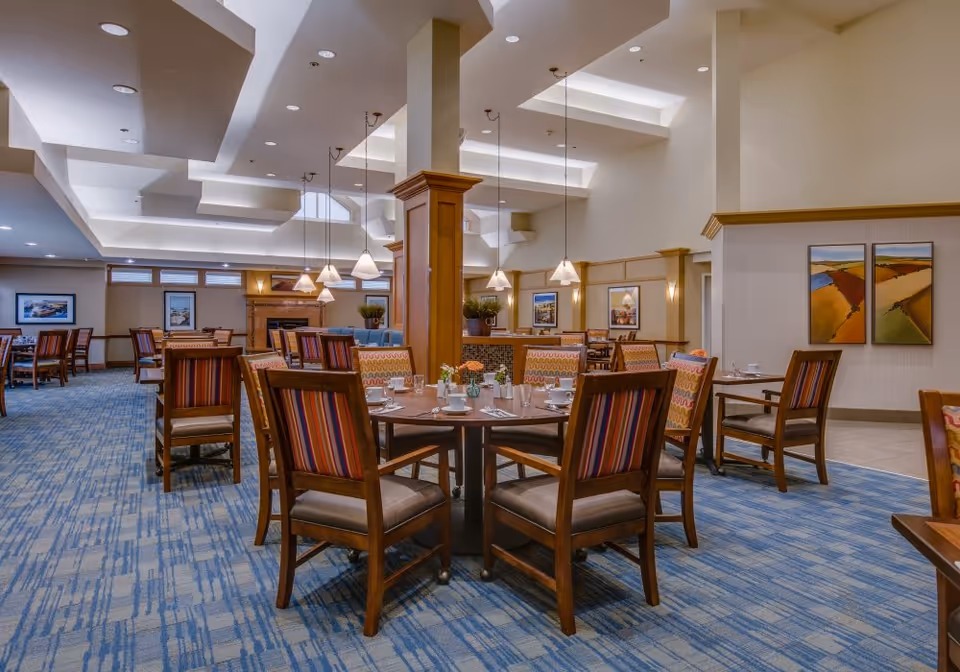 A spacious dining room with multiple wooden tables and chairs featuring striped upholstery. The tables are set with plates, cups, and small flower arrangements. The room has a blue patterned carpet, warm lighting from hanging pendant lamps, and framed artwork on the walls. The ceiling has recessed lighting and architectural details.