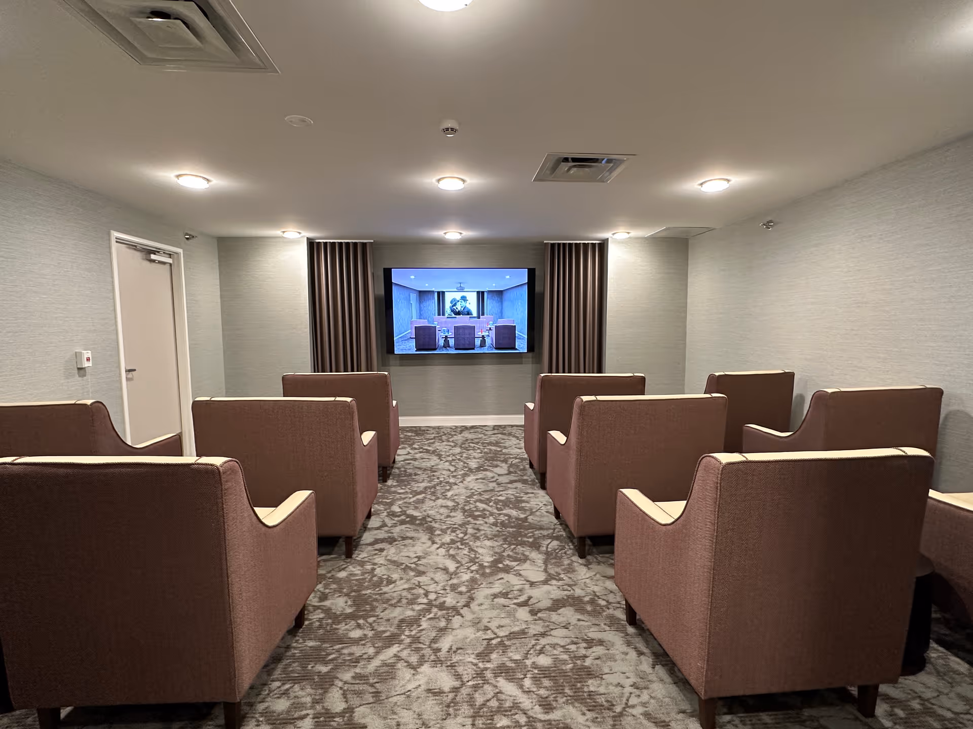 A small theater room with two rows of comfortable armchairs facing a wall-mounted flat screen TV. The room has a carpeted floor, soft lighting from ceiling fixtures, and neutral-colored walls with curtains flanking the TV.