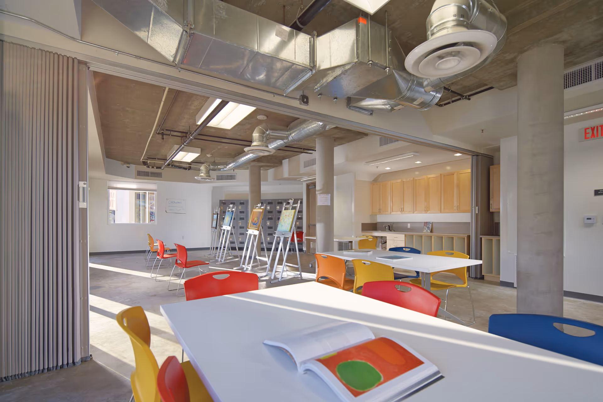 Bright and spacious art room with colorful chairs around white tables, easels displaying artwork, lockers along the back wall, and a kitchenette area with wooden cabinets. The ceiling features exposed ductwork and industrial lighting.