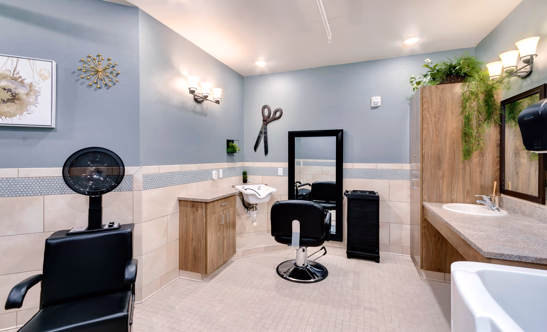 Interior view of a hair salon area in a senior living facility featuring a black salon chair in front of a large mirror, a hair washing station with a sink, a black hair dryer chair, wooden cabinets, a countertop with a sink, decorative wall art, and plants.