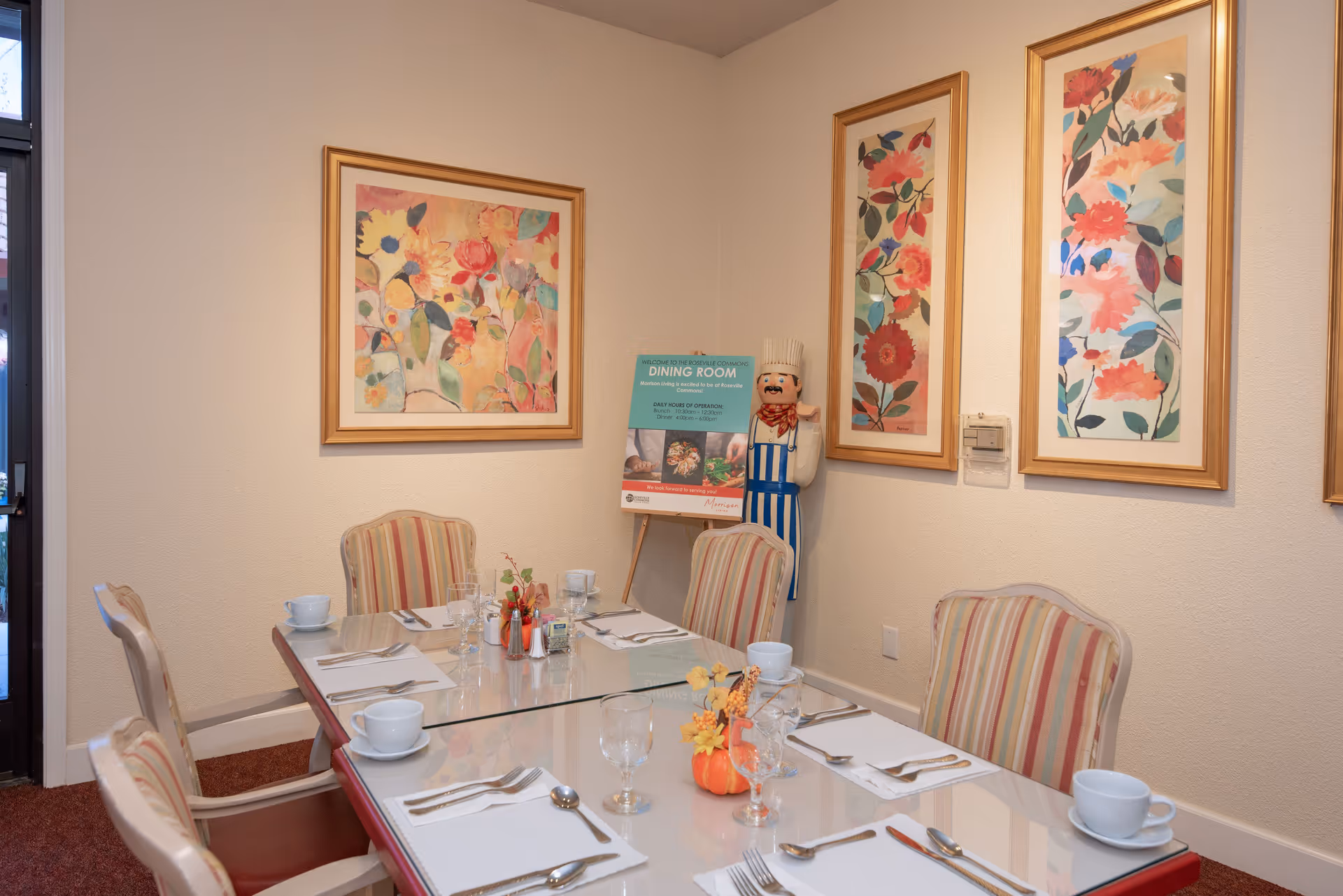 A dining room with a glass-top table set for six people with white cups, glasses, silverware, and napkins. The chairs have striped upholstery. The walls are decorated with three framed floral paintings and a standing chef figurine next to a sign about the dining room.