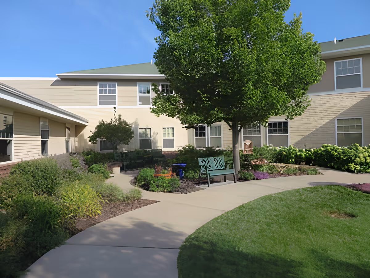 Outdoor courtyard area at Benedictine Living Community- Shakopee Windermere Way featuring a paved walkway, green grass, various shrubs and plants, a large leafy tree, and a green bench. The beige building with multiple windows surrounds the courtyard under a clear blue sky.