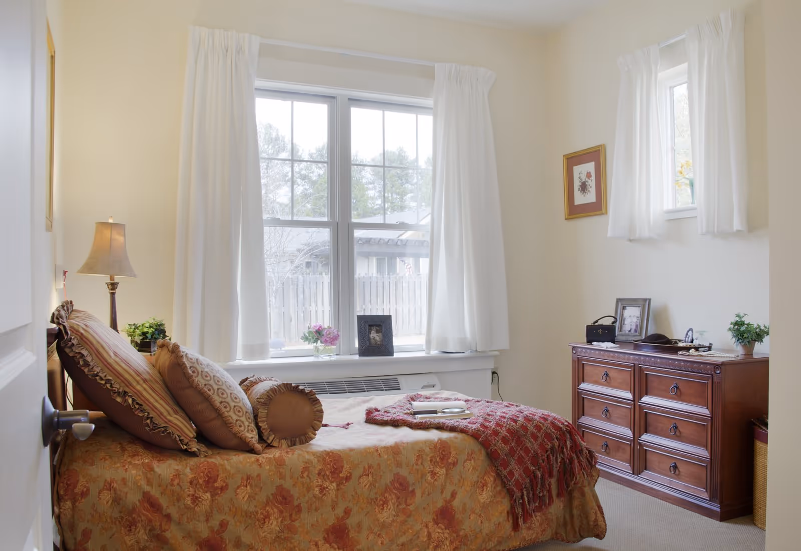 A cozy bedroom with a bed covered in a floral bedspread and multiple pillows. There is a wooden dresser with framed photos and decorative items on top. Two windows with white curtains let in natural light, and a lamp is placed on a small table next to the bed.