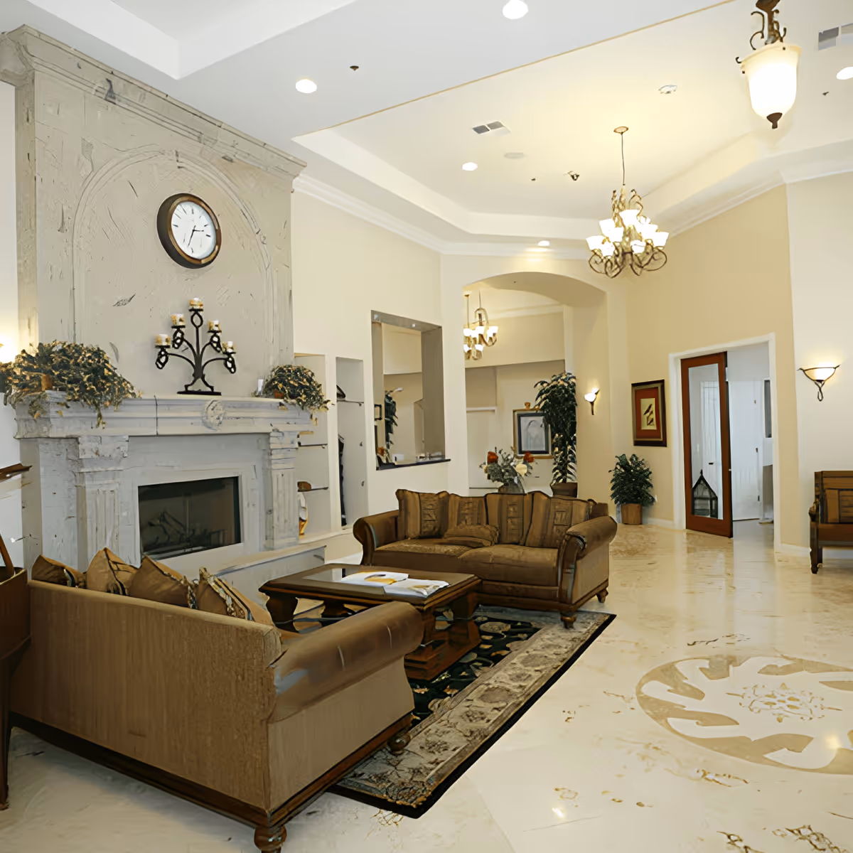 Spacious elegant living room/lobby with sofas arranged around a coffee table, a large fireplace with a clock above it, chandeliers, and decorative plants.