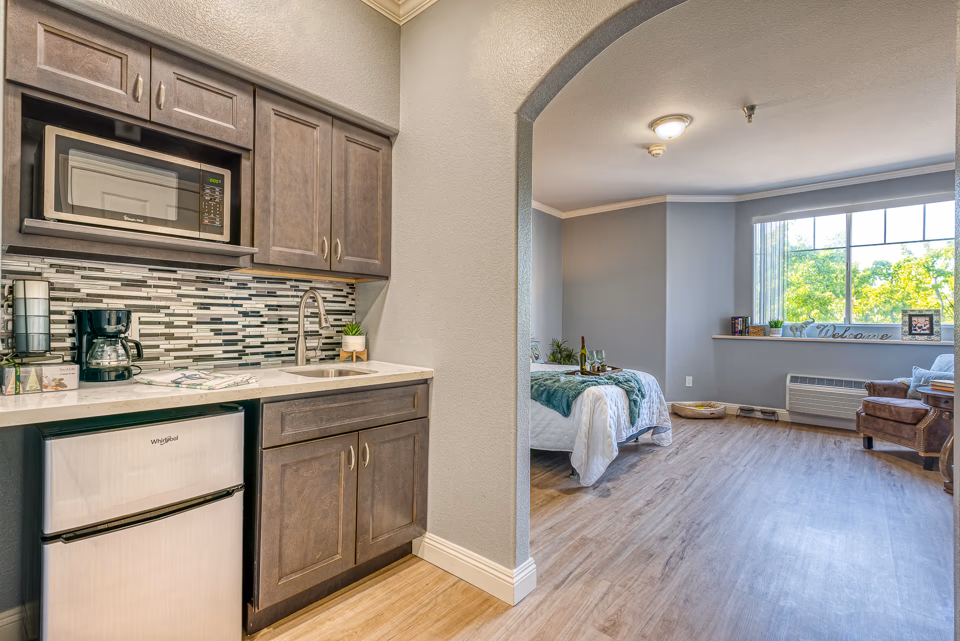 A small kitchenette with microwave, mini-fridge and sink opens through an arched entry into a bright studio bedroom/living area with a bed and large window.