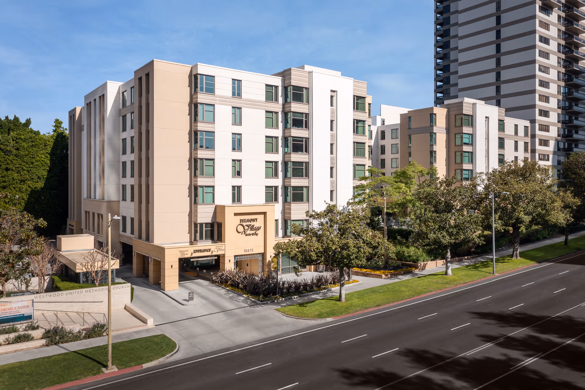 A multi-story Belmont Village senior living building with trees, a sidewalk, and a street.
