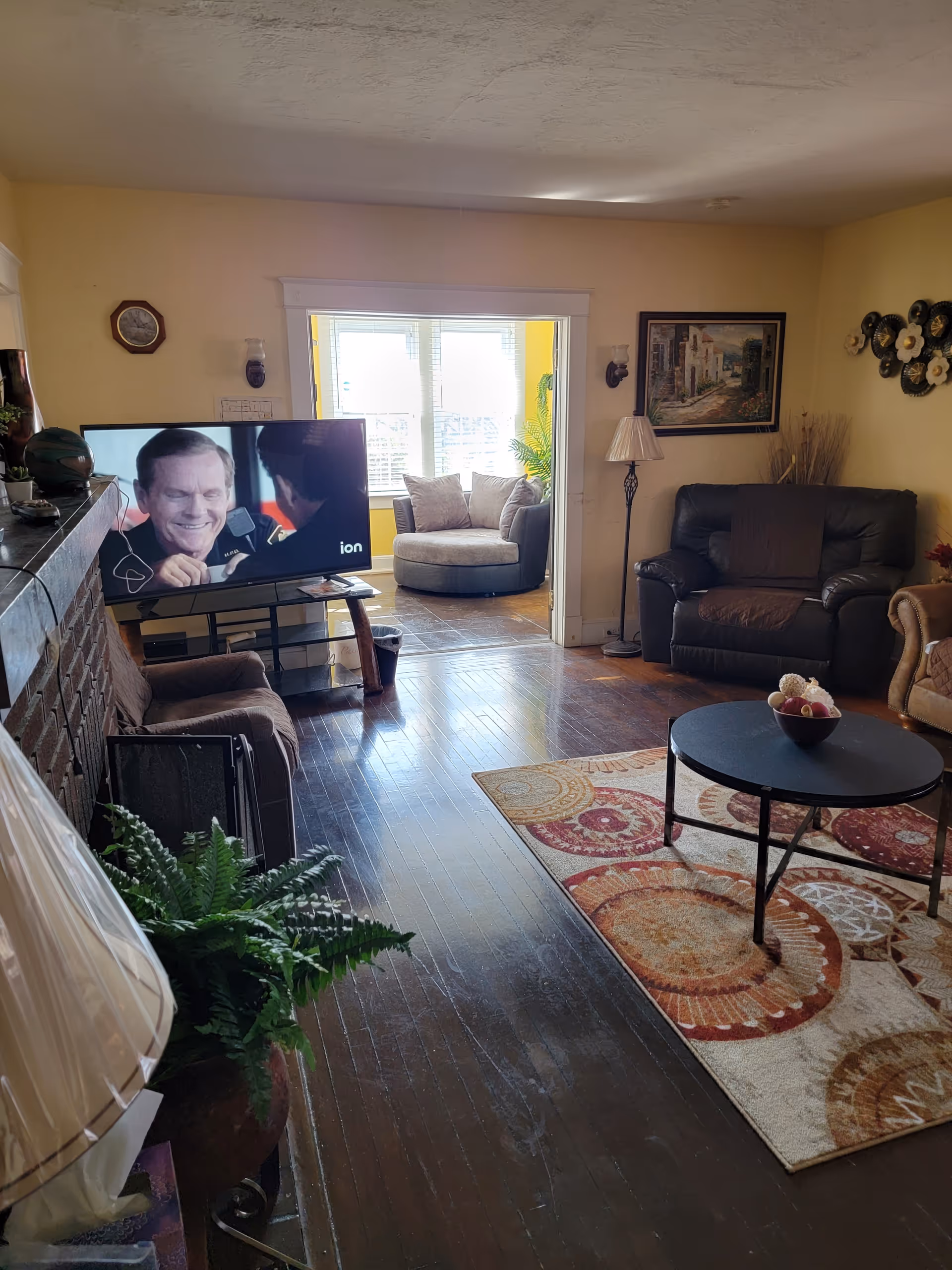 A cozy living room with wooden floors, a patterned area rug, a round black coffee table with a decorative bowl, a brown leather armchair, and a beige armchair. A flat-screen TV is on a stand near a brick fireplace, showing a scene from a TV show. In the background, there is a sunlit sitting area with a round cushioned chair and several pillows. The walls are painted light yellow and decorated with framed artwork and wall sconces.