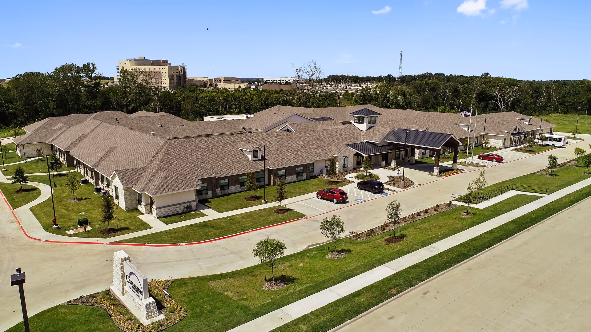 Aerial view of Celebration Senior Living of Denison, a large single-story building with a brown roof and beige stone exterior. The facility is surrounded by well-maintained green lawns, small trees, and paved driveways with parked cars. The entrance features a covered drop-off area with an American flag flying nearby. In the background, there are taller buildings and a wooded area under a clear blue sky.