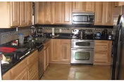 A kitchen with wooden cabinets, black granite countertops, stainless steel appliances including a microwave, oven, dishwasher, and refrigerator. The floor is tiled and there is a decorative tile backsplash.