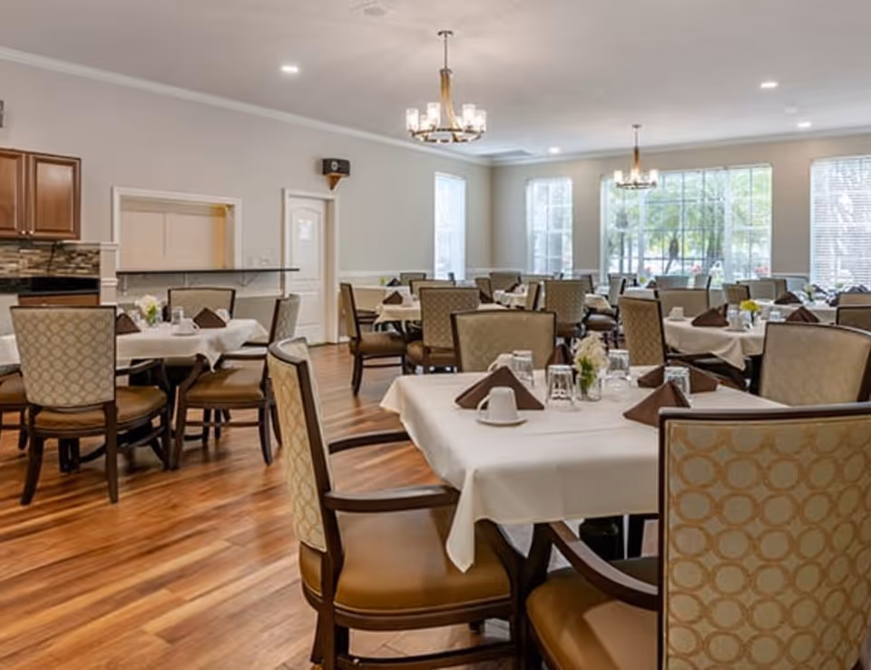 A bright dining room with multiple tables covered in white tablecloths, each set with brown napkins, cups, glasses, and small flower arrangements. The room has wooden flooring, beige walls, large windows letting in natural light, and two chandeliers hanging from the ceiling. There is a kitchen pass-through window and wooden cabinets visible on one side.