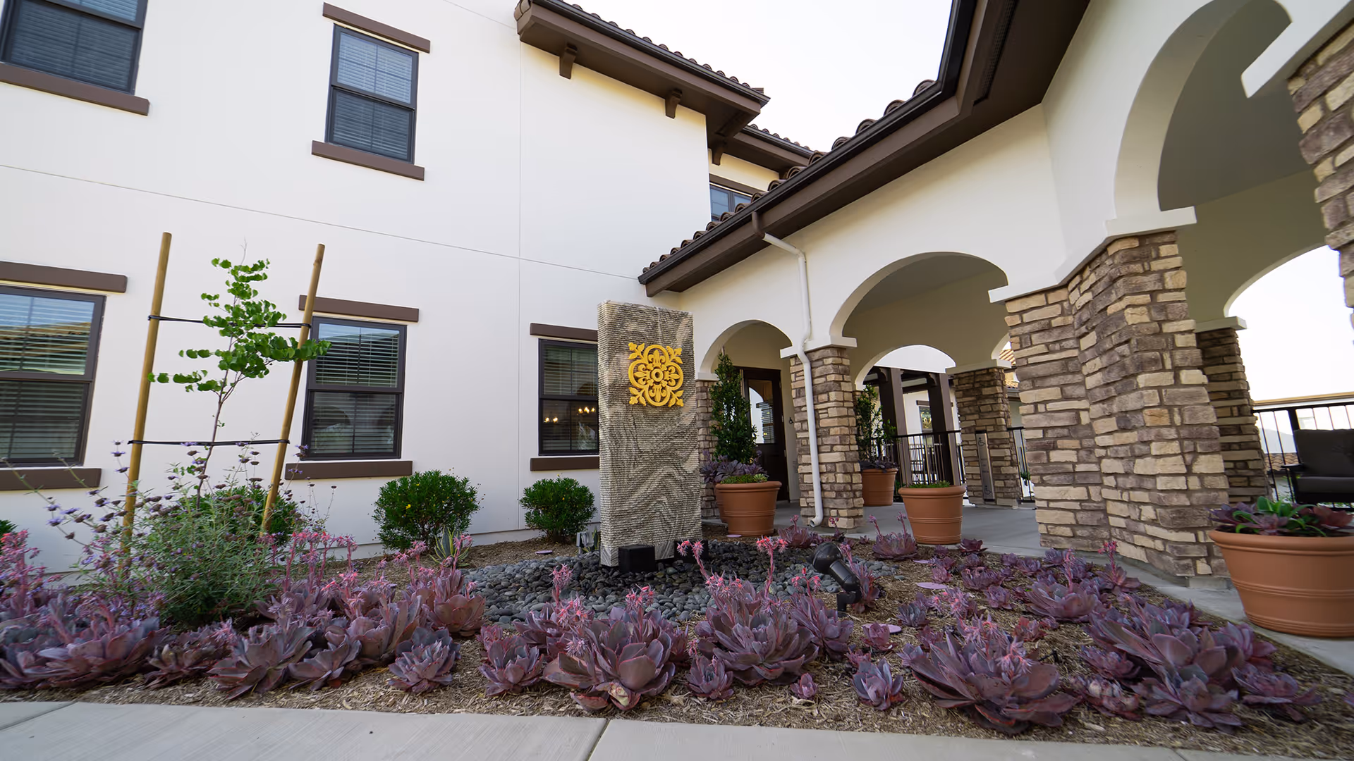 Outdoor view of La Marea Senior Living facility showing a landscaped garden with purple succulents and a stone water feature with a yellow decorative emblem. The building has white walls, brown window frames, and a covered walkway with stone pillars and arches.
