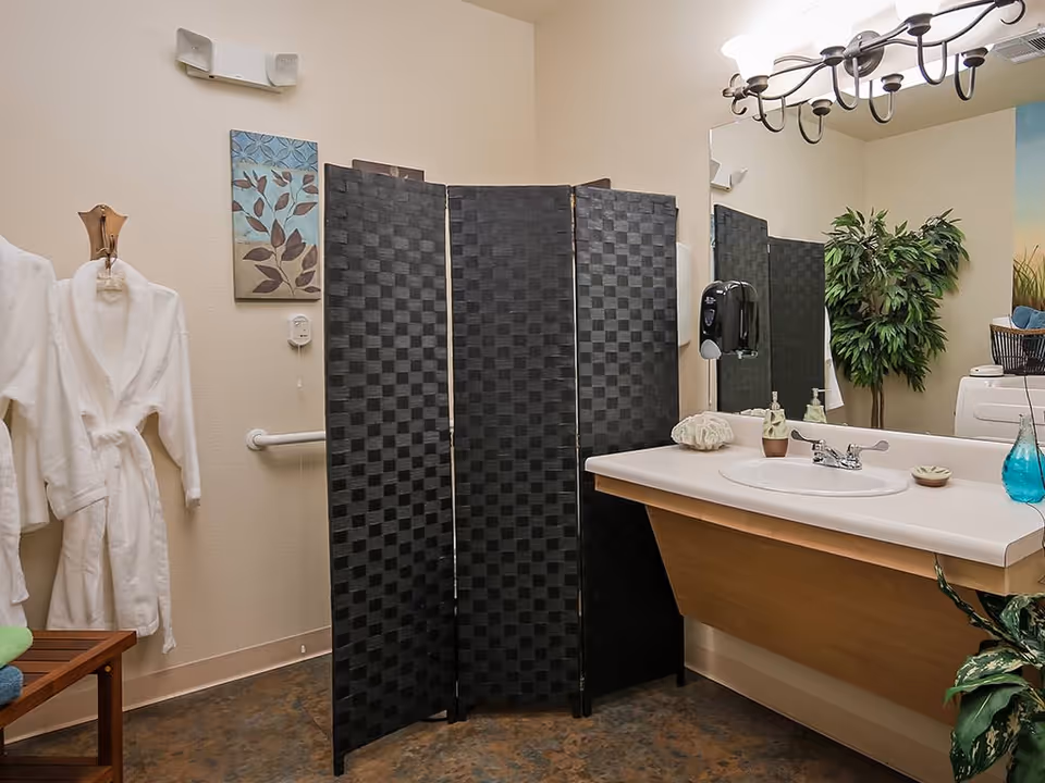 A bathroom with a white sink and countertop, a large mirror above the sink, a black folding privacy screen, two white bathrobes hanging on a wooden hook, a small wooden bench with folded towels, a wall-mounted soap dispenser, decorative wall art, and a potted plant in the corner.