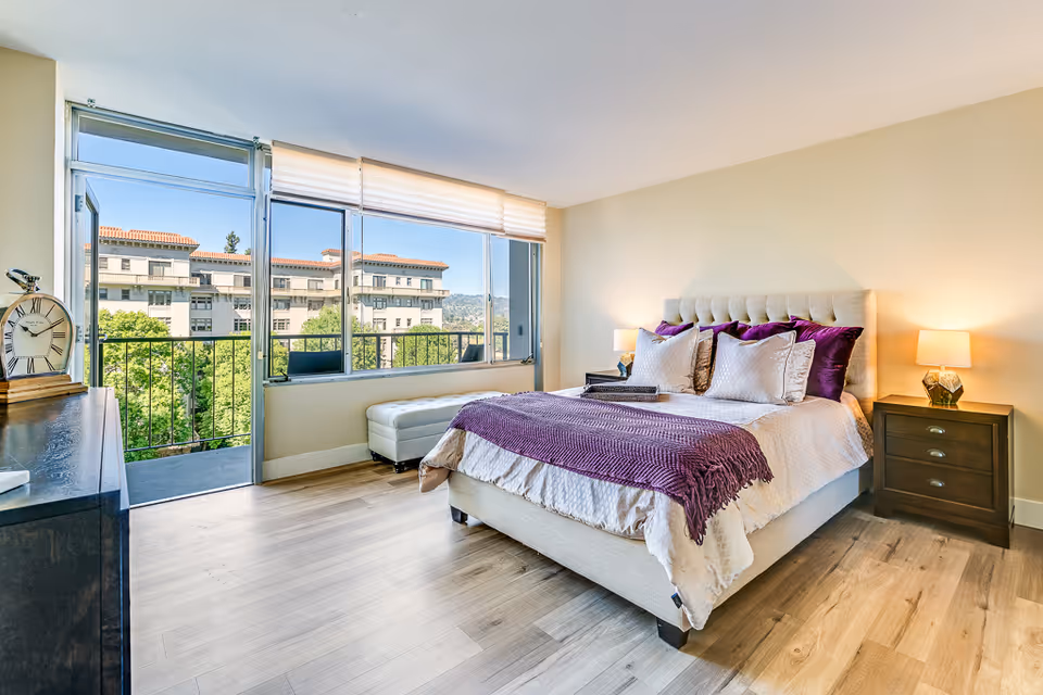 Bright and spacious bedroom with a large bed adorned with white and purple bedding, two bedside tables with lamps, a dresser with a large clock, and a door leading to a balcony with a view of trees and a building outside.