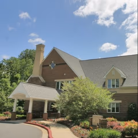 Exterior view of a brick building with a covered entrance, surrounded by landscaped greenery and a clear blue sky with some clouds.