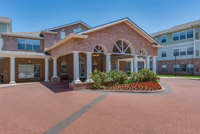 Front exterior view of a senior living facility named Town Village with a brick facade, white columns supporting a covered entrance, landscaped flower beds, and a paved driveway under a clear blue sky.