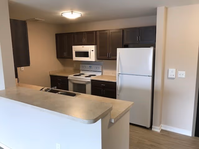 A kitchen area in a senior living facility featuring dark brown cabinets, a white refrigerator, a white electric stove with an oven, a white microwave above the stove, and a countertop with a built-in sink. The walls are painted beige and the floor has light wood-style flooring.
