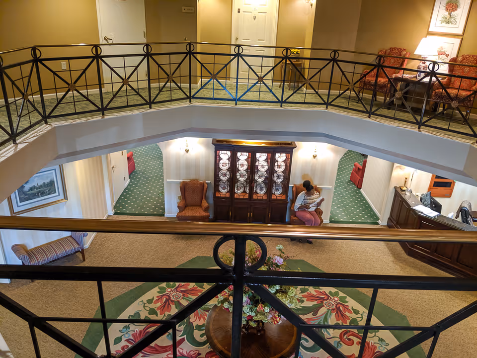 View from an upper floor overlooking a senior living community lobby area with decorative railing, a round table with a floral arrangement on a patterned rug, a wooden cabinet with glass doors displaying plates, two armchairs, and a person sitting reading a newspaper. The area has green and beige carpeting, beige walls, and warm lighting.