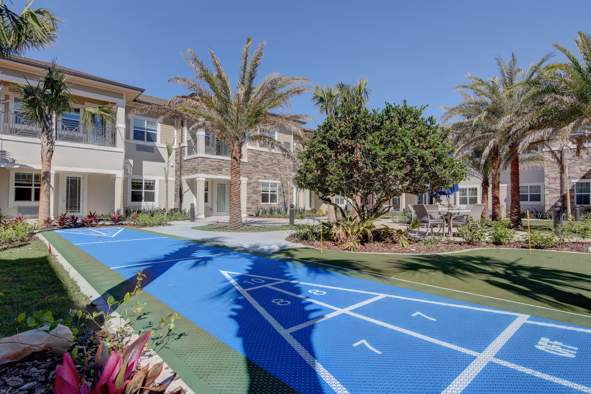 Outdoor courtyard area of Beach House Assisted Living & Memory Care featuring a shuffleboard court, palm trees, landscaped greenery, and patio seating with tables and chairs under a clear blue sky.