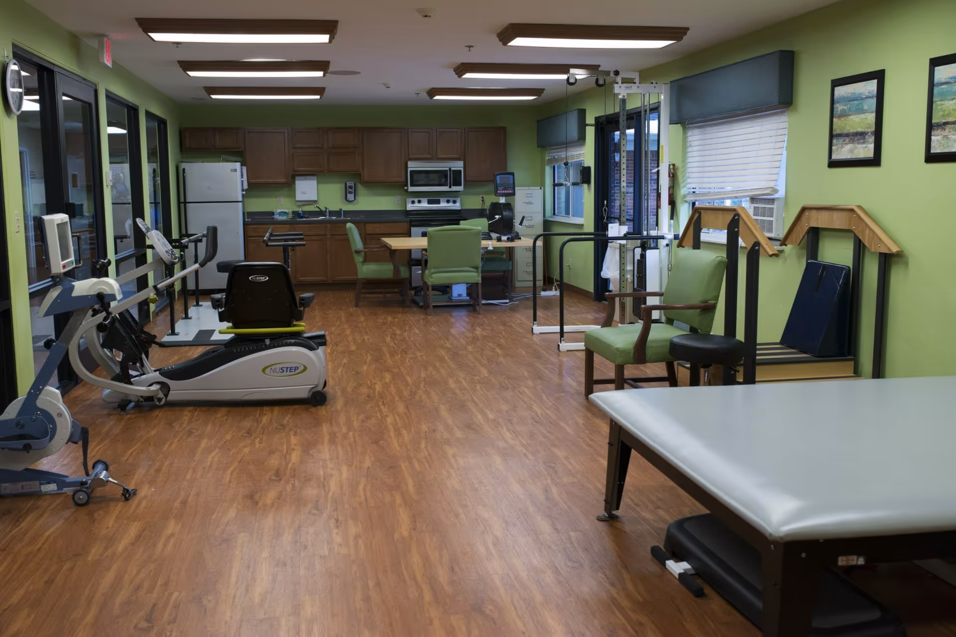 Interior room with green walls and wood flooring featuring exercise equipment including a NuStep recumbent cross trainer, parallel bars, and a therapy table. There is a small kitchenette with wooden cabinets, a refrigerator, microwave, and stove at the back. Green cushioned chairs and framed artwork are also visible.