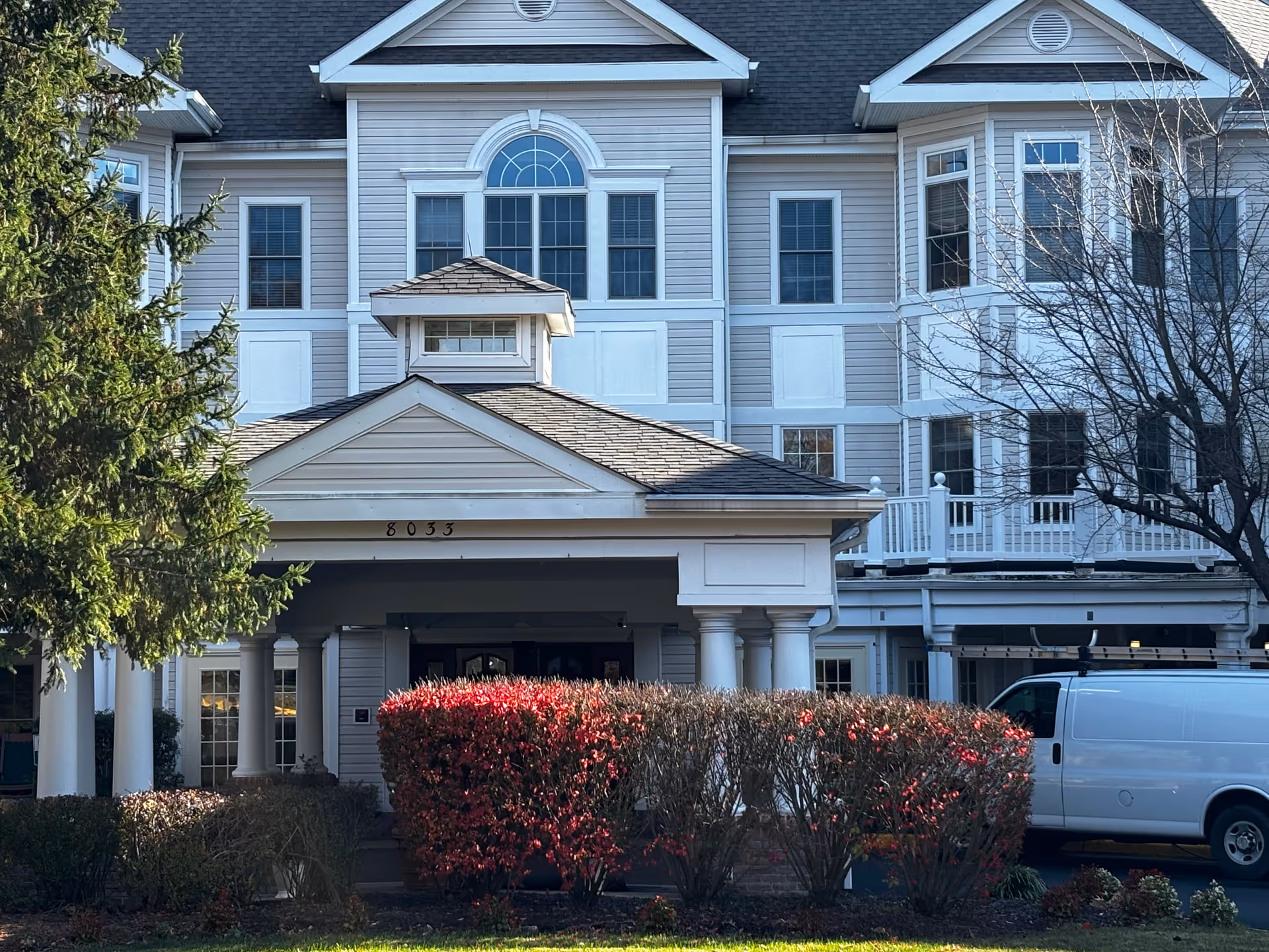 Front entrance of a multi-story white building with a columned portico, red shrubs, and a parked white van.