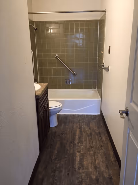 A narrow bathroom with a white bathtub and olive green tiled wall featuring a grab bar, a white toilet, a dark wood vanity with a sink, and a wooden floor. The door to the bathroom is open, showing a silver door handle.