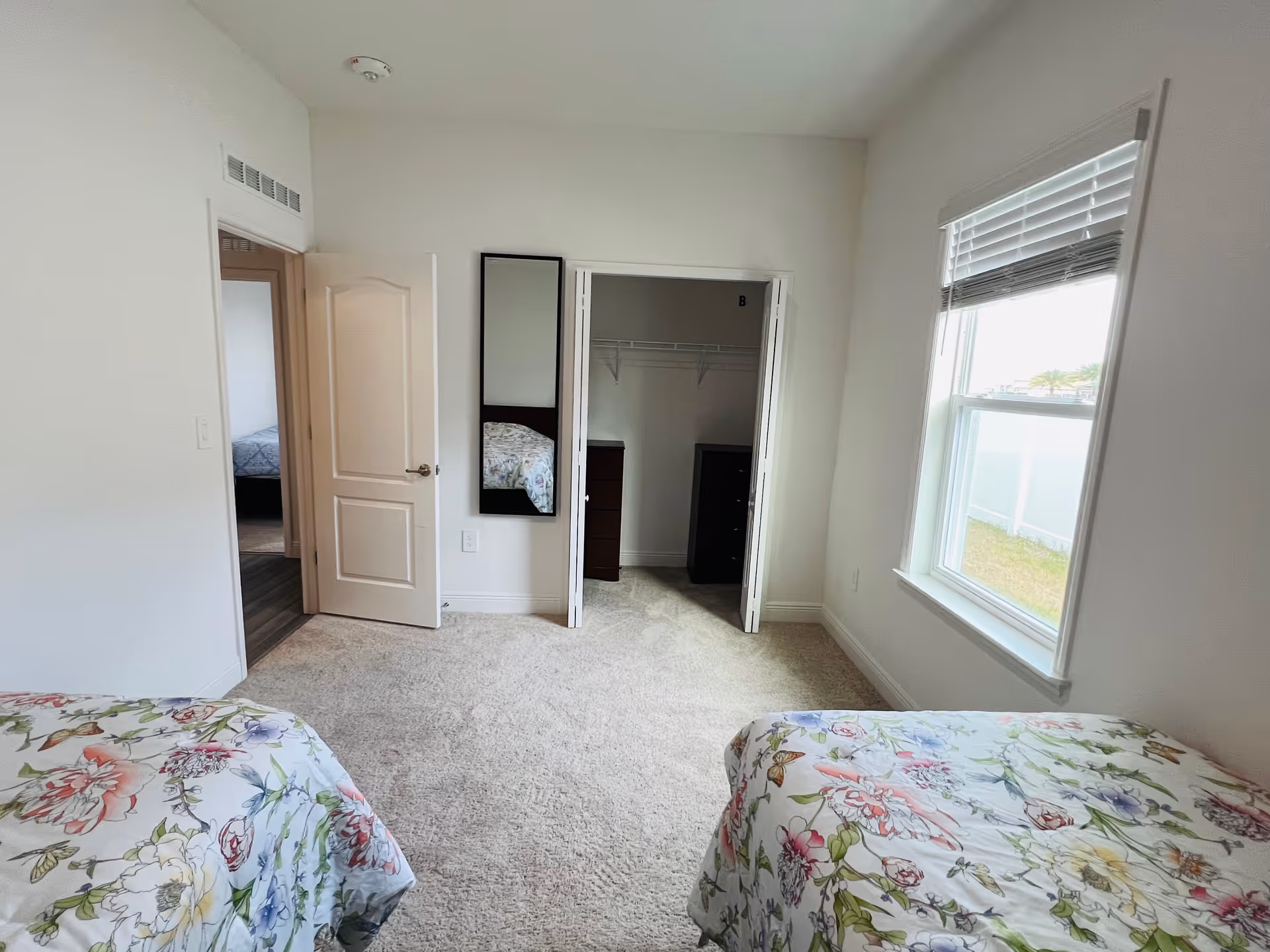 Bright bedroom with two floral twin beds, an open closet with a dresser, a wall mirror, and a window.