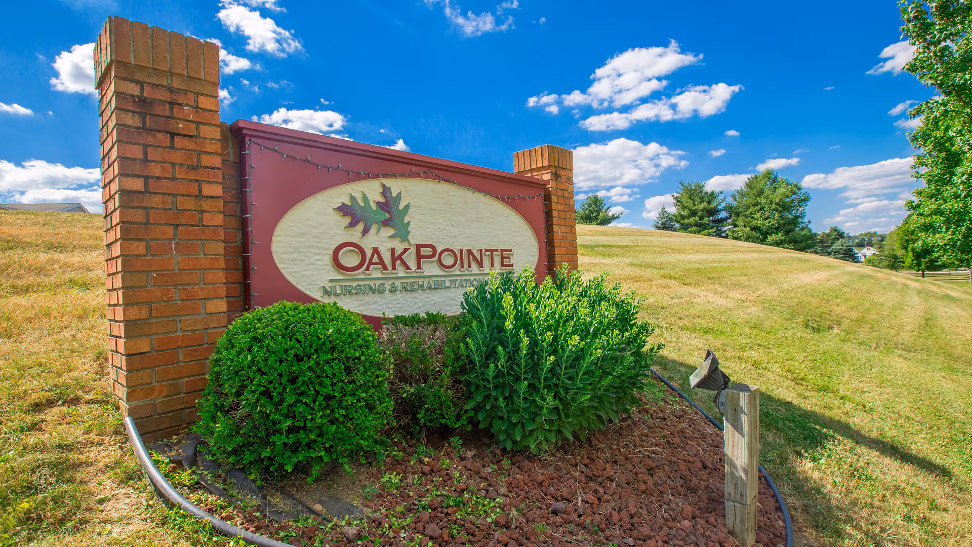 Landscaped outdoor sign for OakPointe Nursing & Rehabilitation set between brick pillars on a grassy hill under a blue sky.