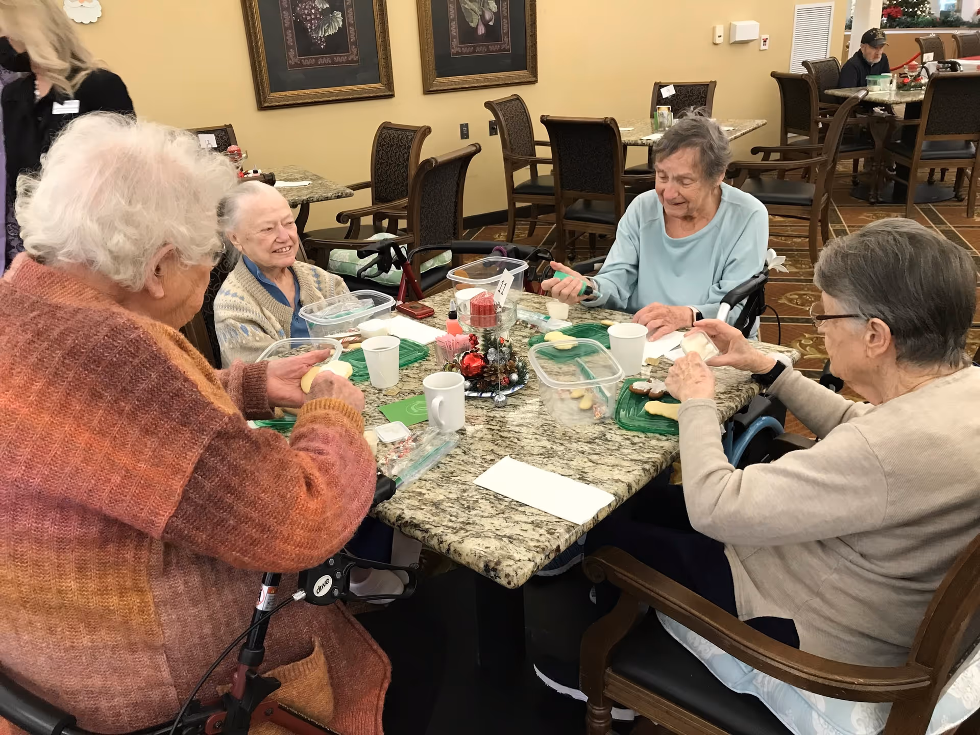 Four elderly women sitting around a granite table in a dining area, engaging in an activity with cookies and icing. The room has framed artwork on the walls and several empty tables and chairs in the background.