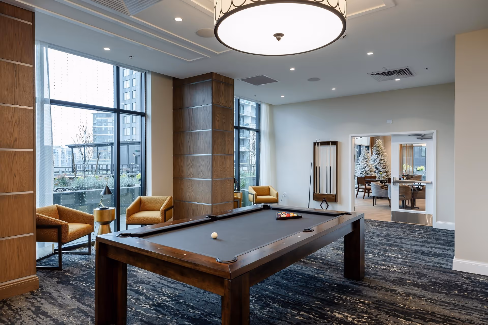 A modern game room featuring a pool table with balls arranged for a game, surrounded by large windows letting in natural light. There are two mustard-colored armchairs with a small round table between them near the windows. The room has a dark patterned carpet, wood-paneled columns, and a large circular ceiling light. Through a doorway, a dining area with a decorated Christmas tree is visible.