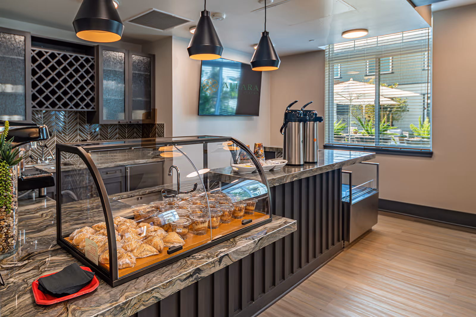 A modern kitchen or snack area in a senior living facility with a marble countertop displaying baked goods and desserts in a glass case. There are three black pendant lights hanging from the ceiling, stainless steel coffee dispensers on the counter, and a window with blinds showing an outdoor patio with umbrellas. Cabinets and a sink are visible in the background.