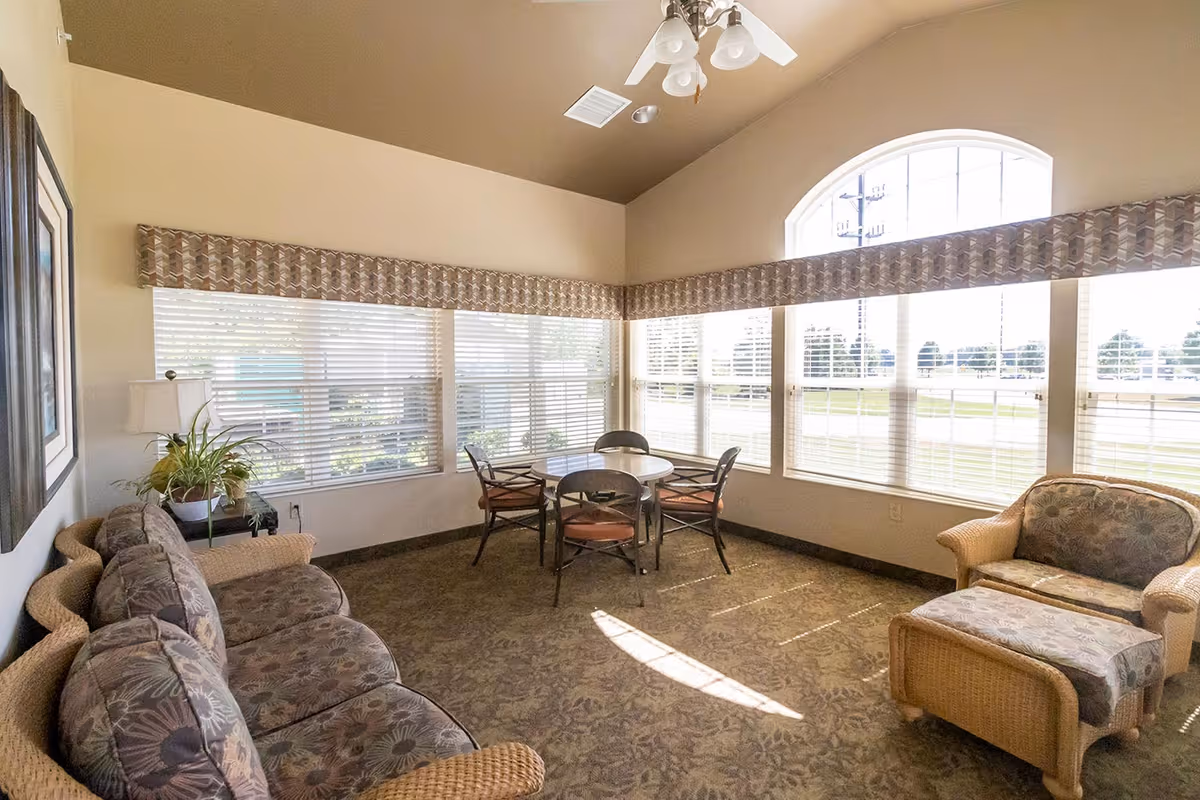 A bright and cozy living room area in an assisted living facility with large windows letting in natural light. The room features a wicker sofa and armchair with floral cushions, a round table with four chairs, a side table with a lamp and a potted plant, and a ceiling fan with lights. The walls are light-colored and the carpet has a floral pattern.