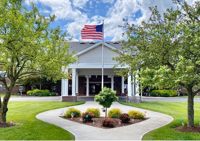 Front exterior view of Canton Regency facility with a large American flag on a flagpole in the center, surrounded by green trees, shrubs, and a well-maintained lawn under a partly cloudy sky.