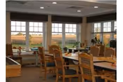 A dining area with several wooden tables and chairs arranged neatly. Large windows line the walls, allowing natural light to fill the room and offering a view of an outdoor field with crops. The room has a warm and inviting atmosphere with neutral-colored walls and ceiling lights.