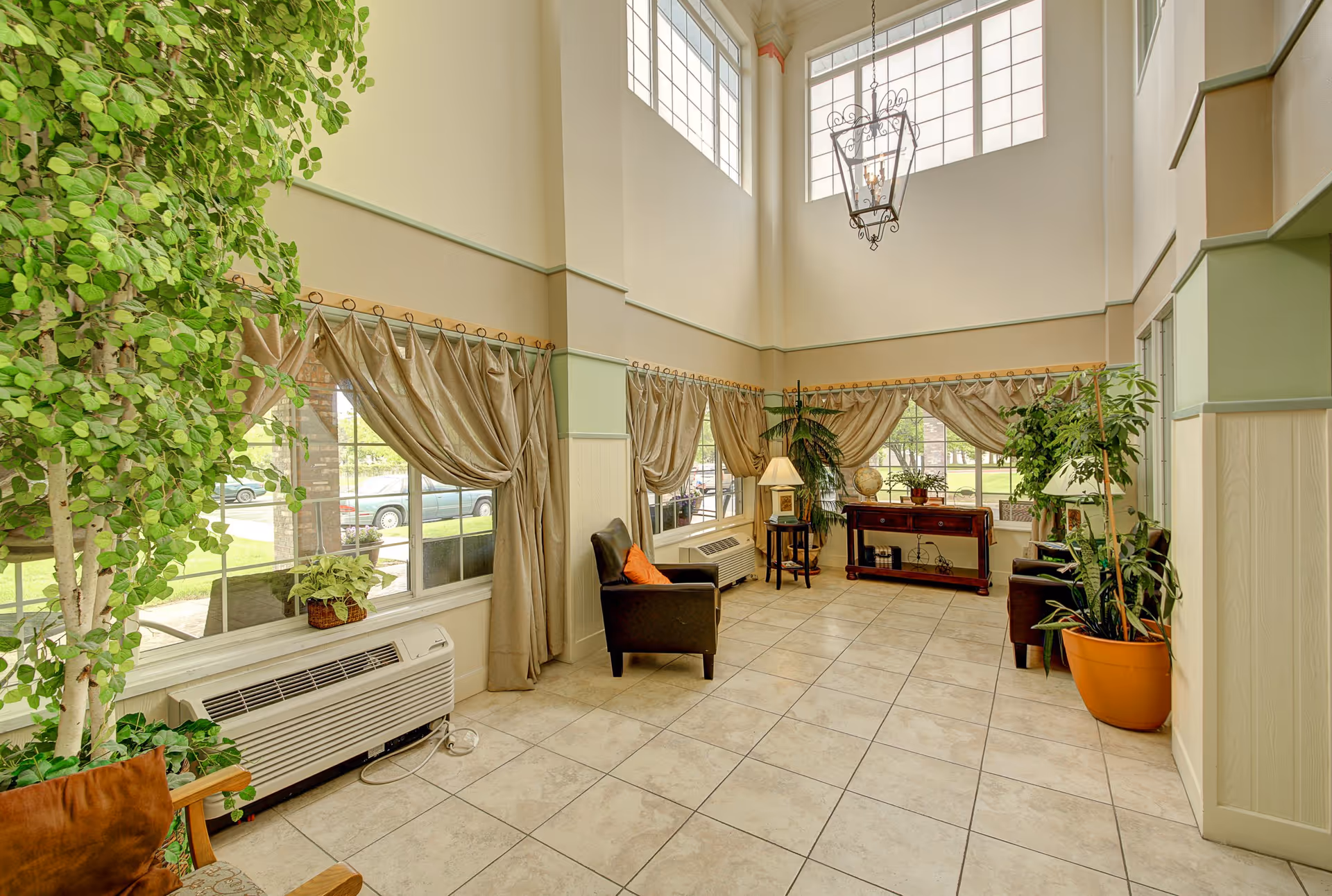 Bright two-story lounge with tiled floor, armchairs, large windows with draped curtains, potted plants and a console table.