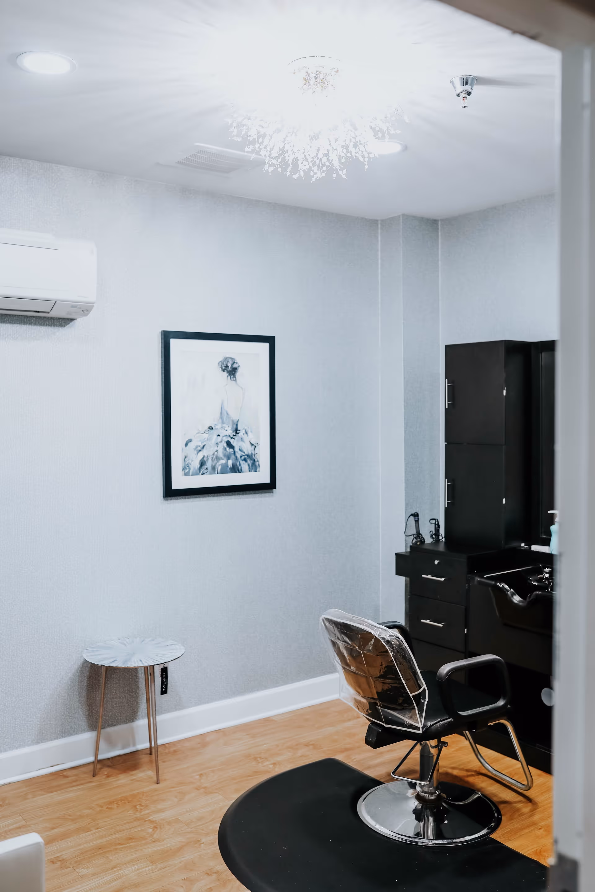 Interior of a small salon or grooming room with a black salon chair covered in plastic, a black cabinet with drawers and a sink, a small round side table, a framed artwork of a woman in a dress on the wall, and a ceiling light fixture.