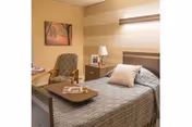 A cozy bedroom in a senior living facility featuring a single bed with a gray patterned bedspread and a pillow. Next to the bed is a wooden nightstand with a lamp and a framed photo. There is a comfortable armchair with a cushion and a small table with magazines. The walls are painted in warm beige tones with a striped accent wall and a framed picture of a tree-lined path.