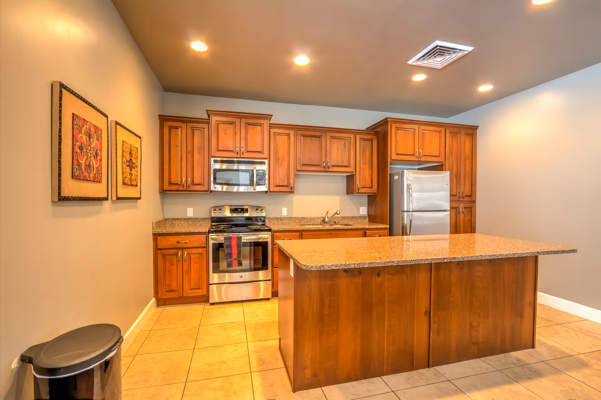 A modern kitchen with wooden cabinets, a stainless steel stove and microwave, a stainless steel refrigerator, a large kitchen island with a granite countertop, beige tiled floor, and two framed artworks on the wall. There is also a black trash can in the corner.