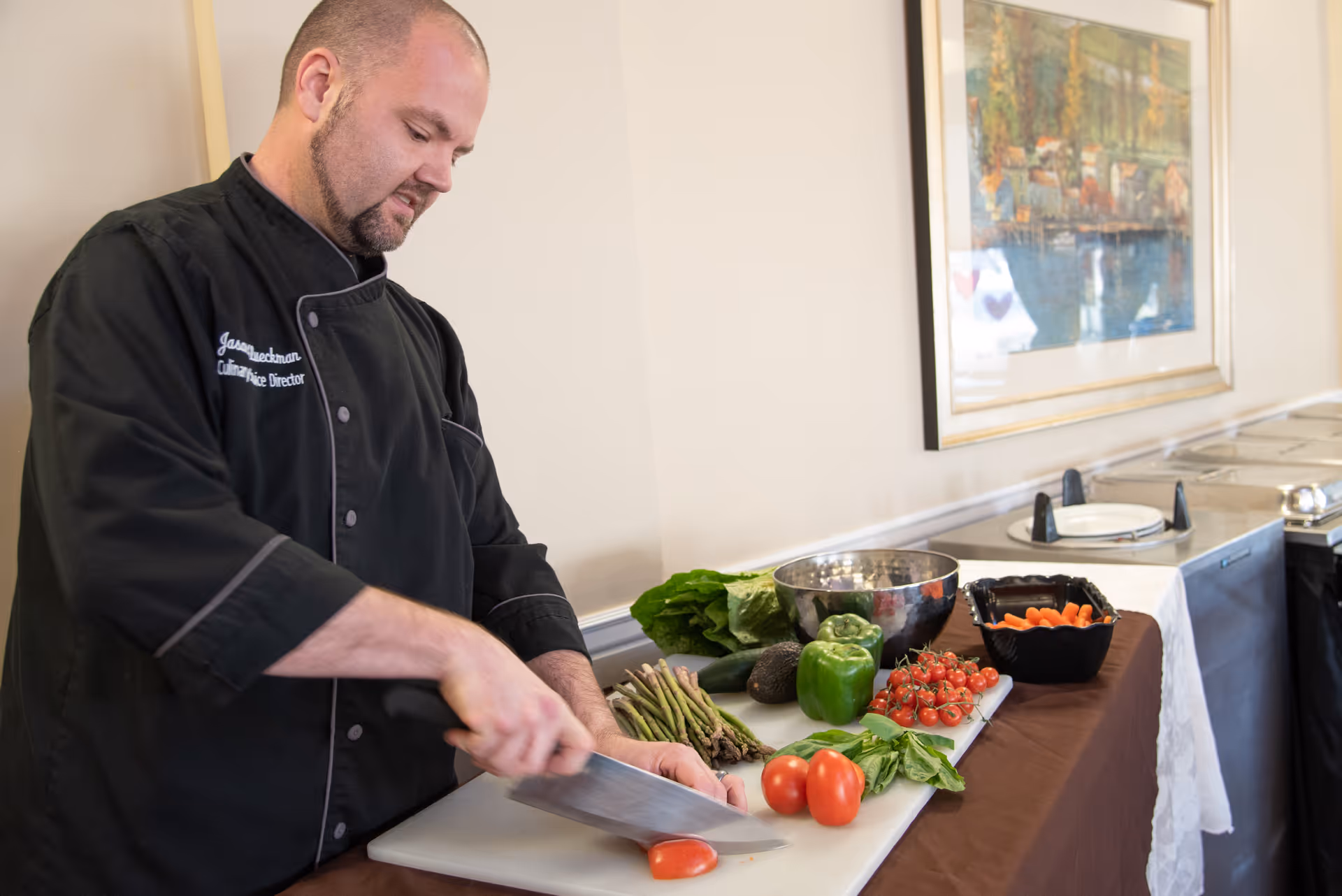 A chef slices a tomato at a prep table surrounded by fresh vegetables and salad bowls next to a buffet line.
