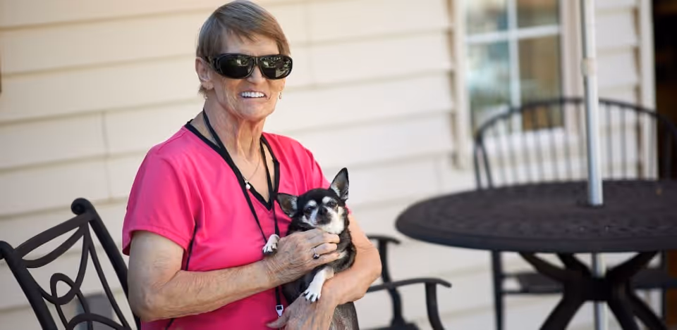 A woman wearing a bright pink shirt and large black sunglasses is sitting outside on a metal chair, holding a small black and white dog. Behind her is a round metal table with an umbrella, and the exterior wall of a building with siding.