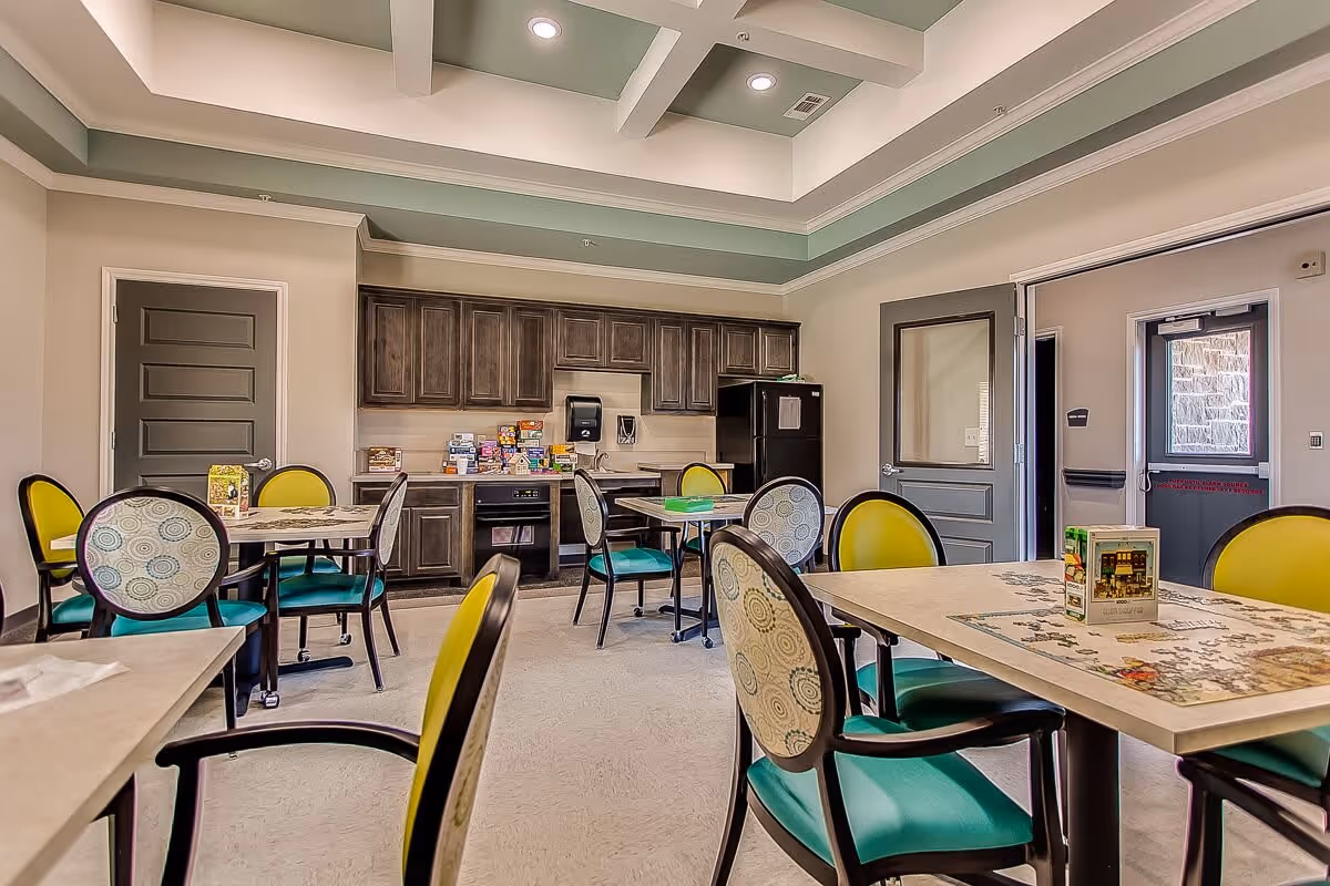 A bright and clean dining room in a senior living facility with several tables and chairs. The chairs have green seats and patterned backs. The room features a kitchenette area with dark wooden cabinets, a black refrigerator, and a countertop with various boxed snacks. The ceiling has a coffered design with recessed lighting. There are two doors, one with a window and another leading outside.
