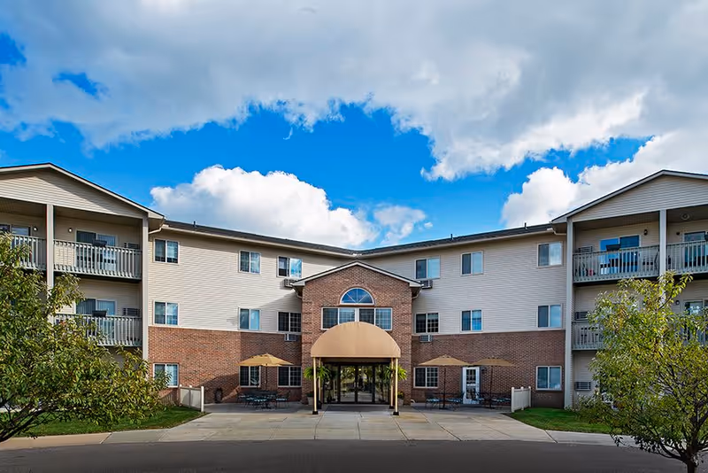 Front exterior view of a three-story senior living facility building with a brick and beige siding facade, balconies on the upper floors, a central entrance with a tan awning, outdoor seating areas with umbrellas, and trees on either side under a partly cloudy blue sky.