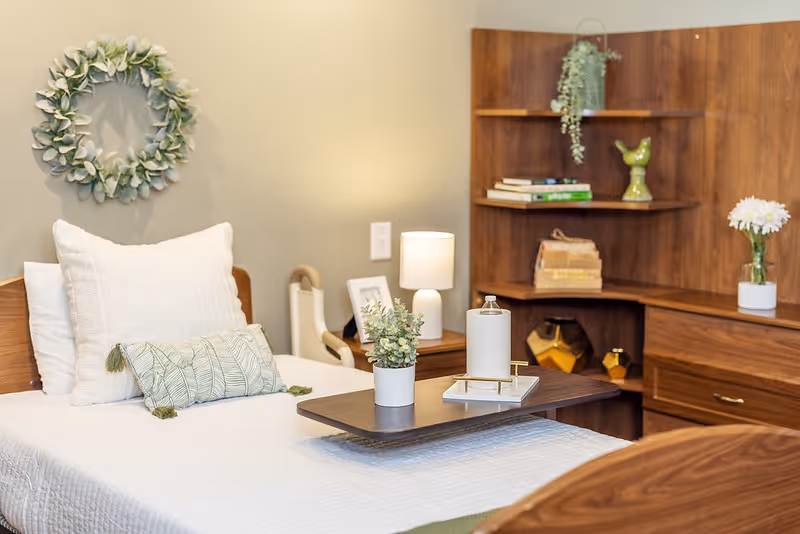 A tidy senior bedroom with a single bed, overbed tray holding a plant and candle, bedside lamp, decorative wreath, and wooden shelving.