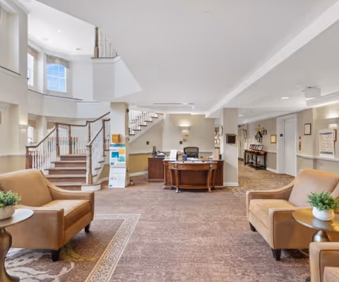 Interior view of a senior living facility lobby with two beige armchairs and small round tables with plants on either side. A wooden reception desk is positioned in the center with office chairs behind it. A staircase with wooden railings leads to an upper floor, and a hallway extends to the right. The space is well-lit with natural light from windows and ceiling lights.