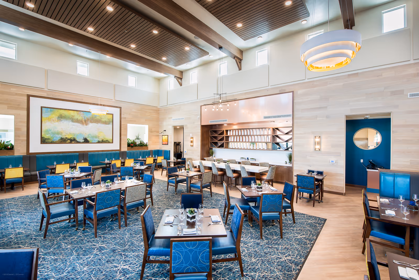 Bright, modern dining room with multiple set tables and blue upholstered chairs beneath high ceilings and decorative lighting.
