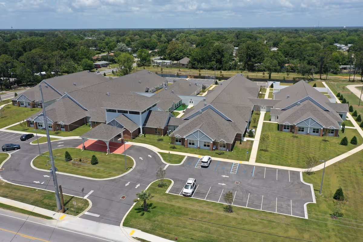 Aerial view of Bay Port of Fort Walton Beach facility showing multiple connected buildings with gray roofs, surrounded by green lawns, parking lots with a few cars, and paved roads with marked lanes and sidewalks. Trees and residential houses are visible in the background under a partly cloudy sky.