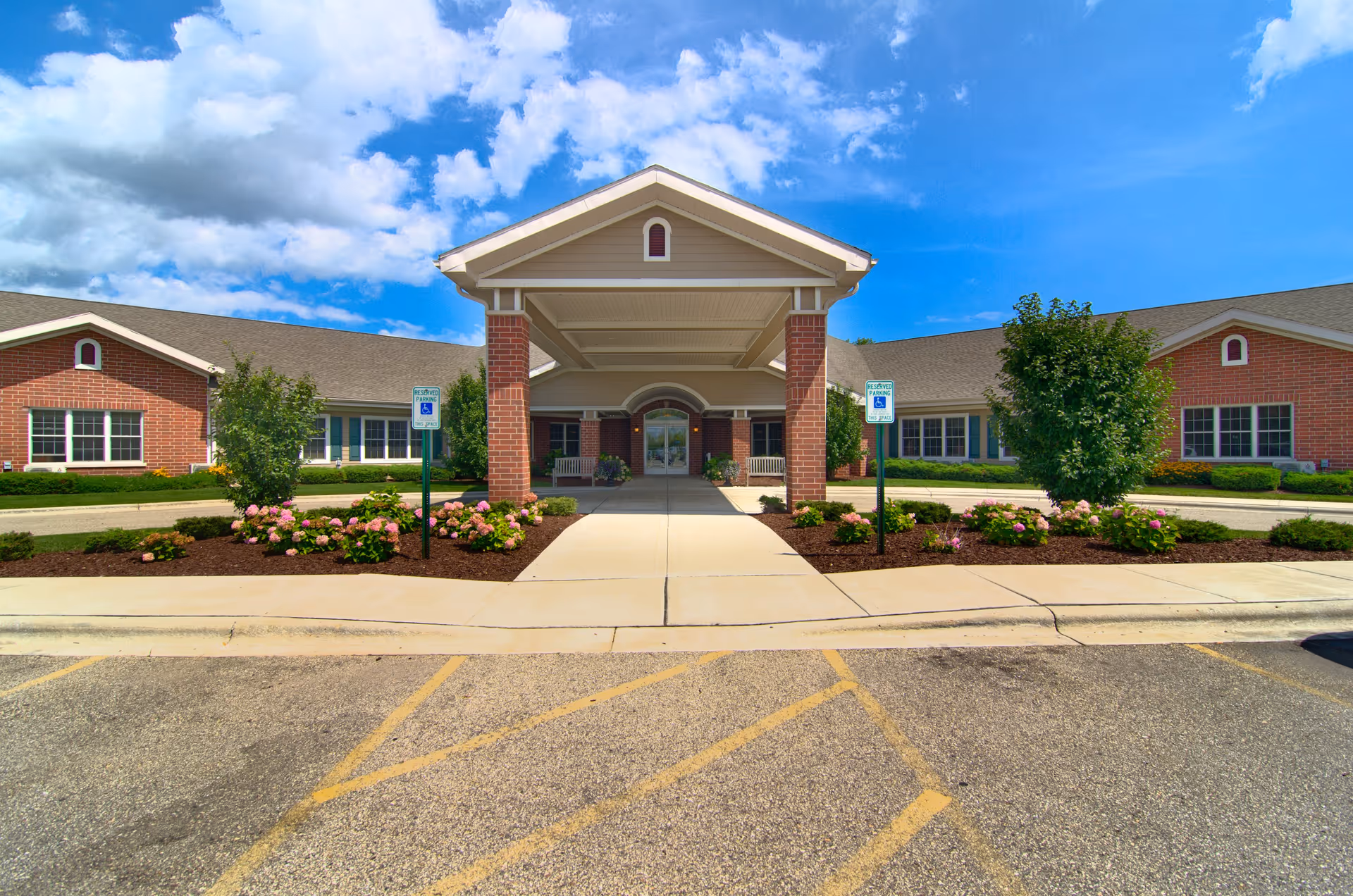 Front entrance of Parkside Manor Assisted Living and Memory Care facility with a covered walkway supported by brick columns, landscaped flower beds, and a clear blue sky with some clouds.