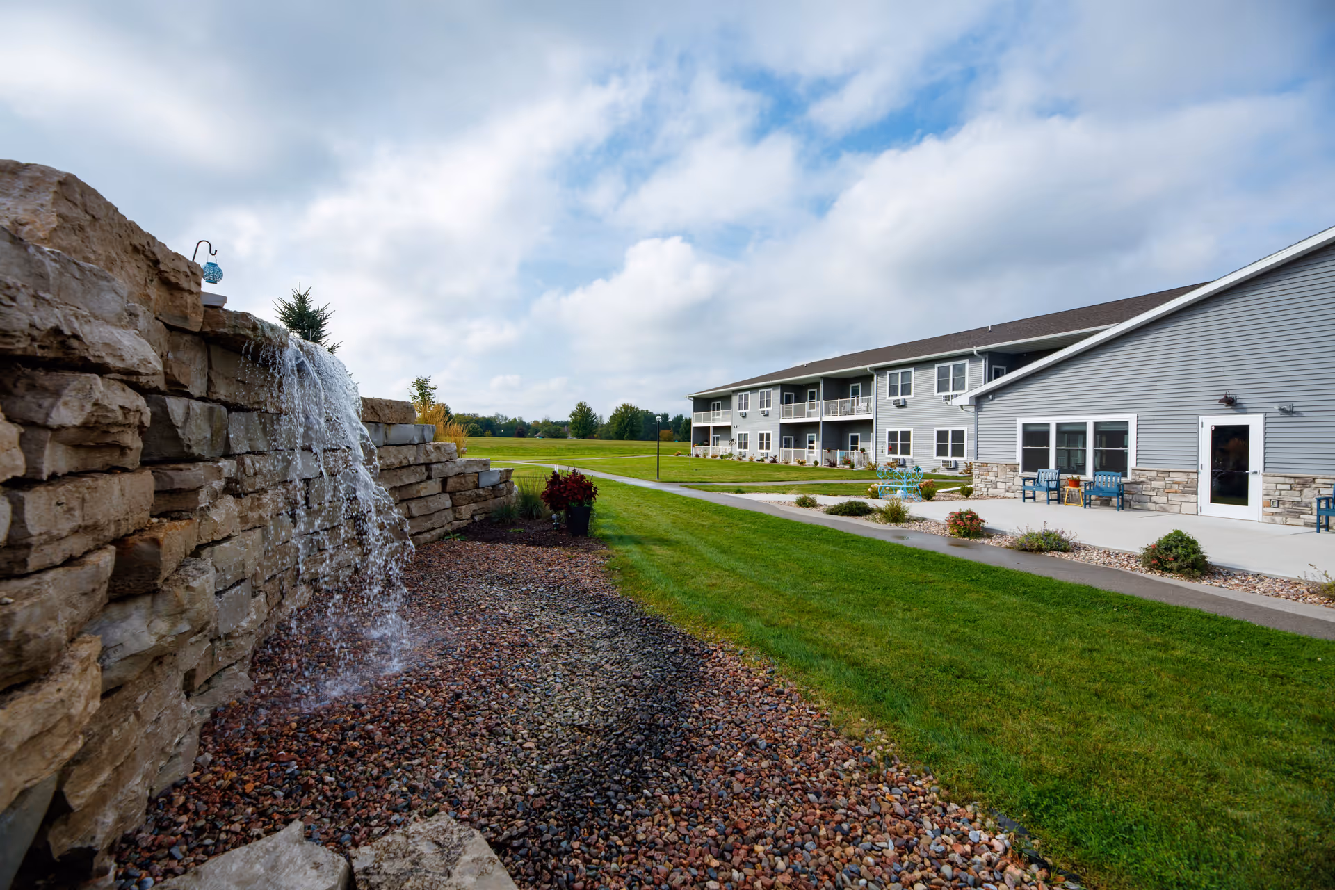 Exterior view of a senior living building with balconies and a patio, a green lawn, and a stone waterfall feature in the foreground.
