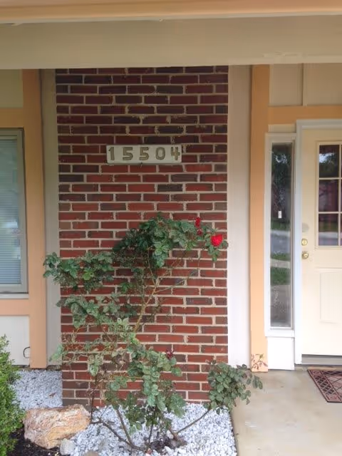 Front entrance with a brick pillar displaying the house number 15504, a rose bush in front, and a beige entry door.