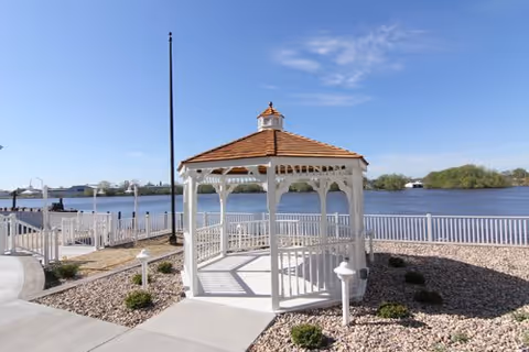 White wooden gazebo with a brown shingled roof situated on a paved area next to a body of water, surrounded by a white fence and landscaped with small bushes and rocks under a clear blue sky.