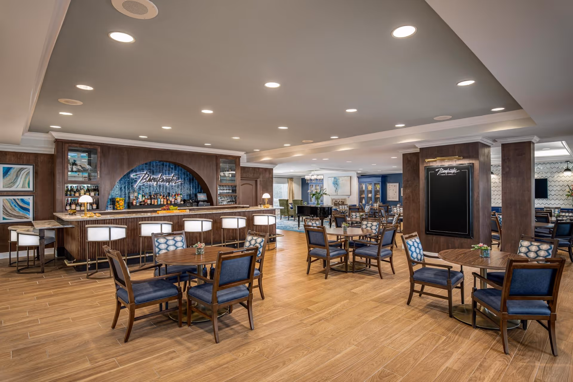 A spacious and well-lit dining area in a senior living facility named Mangrove Bay, featuring multiple round wooden tables with blue cushioned chairs. In the background, there is a bar area with a neon sign that reads 'Blackwater' and several bar stools. The room has wooden flooring, recessed ceiling lights, and decorative artwork on the walls.